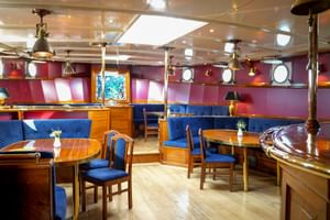 Interior of Mare Frisium ship lounge with wooden tables, blue upholstered seating, brass pillars, pendant lights, and round portholes on red walls.