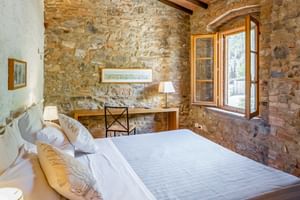Rustic double room with exposed stone walls, wooden beams, white bedding, desk with lamp, and arched window overlooking countryside.