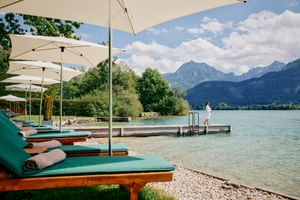 Wooden sun loungers with green cushions and white umbrellas line a pebble beach. A wooden dock extends into a turquoise lake with mountains beyond.