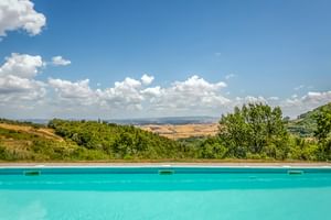 Turquoise pool at Le Valli Agriturismo in Casciana Terme with panoramic view of Tuscan hills under blue sky with white clouds.