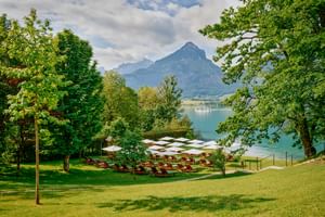 Lakeside garden at Landhaus zu Appesbach with sun loungers and white parasols on green lawn, overlooking turquoise lake and mountains.