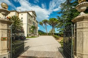 Offene schmiedeeiserne Tore mit Steinsäulen führen zur Villa Soligo. Ein Kiesweg erstreckt sich zum weißen Gebäude mit grünen Fensterläden.