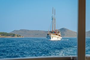 White sailing vessel MS Macek with two masts cruising on blue water, with rocky coastal islands and hills in the background under clear sky.