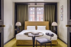 Modernes Doppelzimmer im The Dean München mit senfgelbem Bettgestell, weißer Bettwäsche, olivfarbenen Vorhängen, rosa Tischlampen und gelbem Teppich.