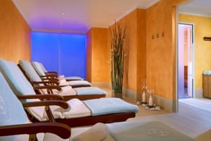 Modern spa relaxation room at Sheraton Essen with wooden lounge chairs, orange walls, blue ambient lighting, and decorative bamboo plants.