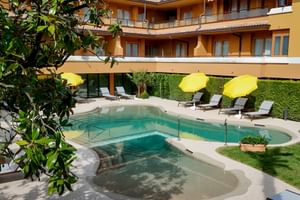 Curved pool with shallow entry at Du Lac et Bellevue hotel in Bardolino. Yellow umbrellas and loungers line the pool, backed by hedge and orange hotel building.