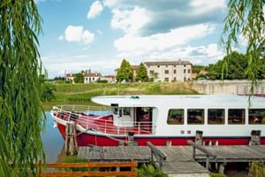 Rot-weißes Flusskreuzfahrtschiff MS Ave Maria am Holzsteg. Weidenbäume rahmen die Szene mit italienischen Landhäusern im Hintergrund.