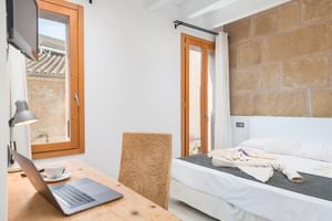 Modern hotel room with natural stone wall, white bed, wooden desk with laptop, wicker chair, and French doors opening to balcony.