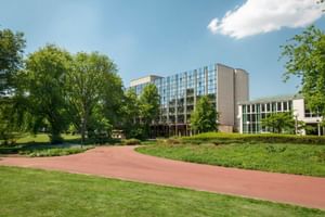 Modern Sheraton Essen hotel building with glass facade surrounded by green parkland, trees and a red brick pathway under blue sky.