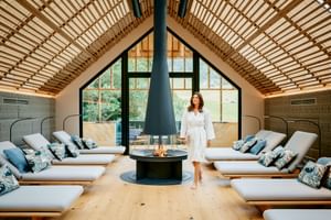 Spa relaxation room with wooden barrel ceiling, central fireplace, and loungers arranged symmetrically. Woman in bathrobe stands by the fire.