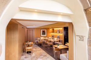 Moderner Frühstückssaal im Hotel Le Colombier mit Holztischen, beigen Stühlen, warmer Beleuchtung und Steinbogen-Eingang in Colmar.