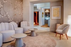 Elegant spa lounge with white curved chairs, round tables on beige carpet, floral wall art, and view through archway to sauna entrance.