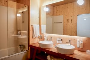 Bright bathroom with wood paneling, two round vessel sinks on wooden vanity, backlit mirror, glass shower enclosure, and modern fixtures.