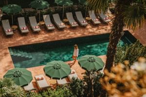 Outdoor pool at Parkhotel Mondschein Bozen with green umbrellas, sun loungers, and palm trees. A woman walks along the turquoise pool.