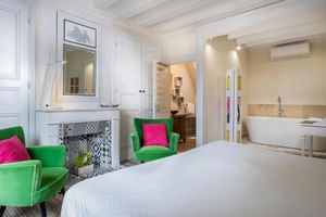 Elegantes Hotelzimmer mit weißem Bett, grünen Sesseln mit rosa Kissen, dekorativem Kamin und offenem Bad mit freistehender Badewanne.