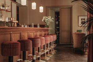 Elegant hotel bar with wooden counter, red velvet bar stools, white pendant lights, and palm plants at Parkhotel Mondschein Bozen.