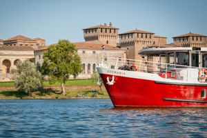 Rot-weißes Passagierschiff MS Ave Maria auf blauem Wasser mit historischen Festungsmauern und Türmen in Mantua, Italien unter klarem Himmel.