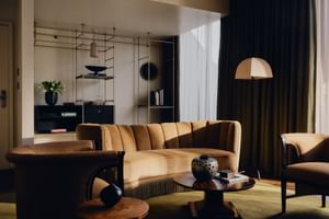 Elegante Lounge im The Dean Munich Hotel mit geschwungenem Samtsofa, Sesseln, rundem Couchtisch, Stehlampe und modernem Regal.