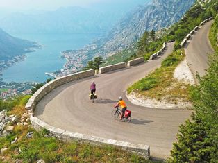 Two cyclists on a winding road with a view of the Bay of Kotor surrounded by mountains