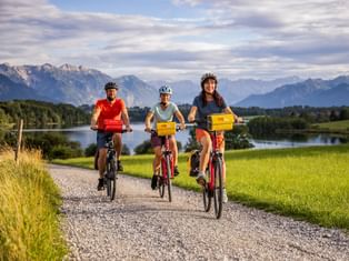 Three cyclists riding on a gravel path near Riegsee in Bavaria, with mountains, lake, and green meadows in the background under a cloudy sky.