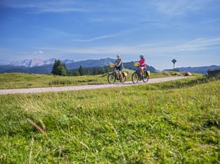Two cyclists on e-bikes riding along a paved path through green meadows in Chiemgau, with mountain ranges visible in the background.