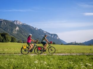 Zwei Radfahrer fahren durch grüne Wiesen im Chiemgau mit Schloss Hohenaschau und Bergen im Hintergrund unter blauem Himmel.