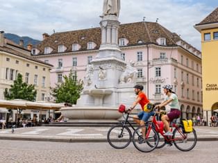 Zwei Radfahrer fahren am weißen Monumentbrunnen am Waltherplatz in Bozen vorbei, mit historischen pastellfarbenen Gebäuden und Straßencafés.