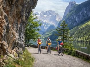 Three cyclists on lakeside path at Gosausee with Dachstein mountain range in background. Rocky cliff on left, alpine lake and forested slopes visible.