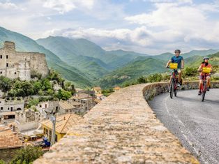 Two cyclists on a road, a ruined castle and wooded mountains in the background