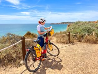 Cyclist with helmet reading a map on a touring bike with yellow panniers, standing on a coastal path overlooking turquoise ocean in Algarve.