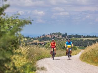Two cyclists on a gravel path in Val d'Orcia, Tuscany. A hilltop village with cypress trees is visible in the background under a blue sky.