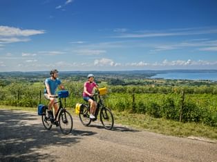 Two cyclists with yellow panniers on a road near Cavaion Veronese, overlooking Lake Garda with vineyards and mountains in the background.