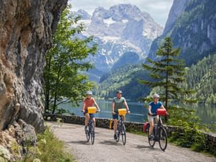 Three cyclists riding along a lakeside path with steep cliffs on the left and a calm lake reflecting snow-capped mountains in the background.