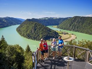 Four people on a viewing platform overlooking the Schlögener Schlinge, a dramatic horseshoe bend of the Danube River surrounded by green hills.