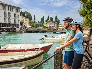 Two cyclists with helmets standing at waterfront in Peschiera, Lake Garda. Boats moored in turquoise water, buildings and cypress trees in background.