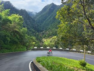 Radfahrer in roter Jacke auf kurvenreicher Bergstraße nahe Gambao, Madeira, mit grünem Tal und dramatischen Berggipfeln im Hintergrund.