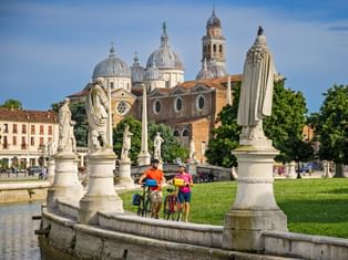 Two cyclists with bikes standing by statues at Prato della Valle in Padua, with the Basilica of Santa Giustina visible in the background.