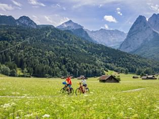Two cyclists surrounded by flower meadows and alpine huts, with the Zugspitze massif near Garmisch-Partenkirchen in the background