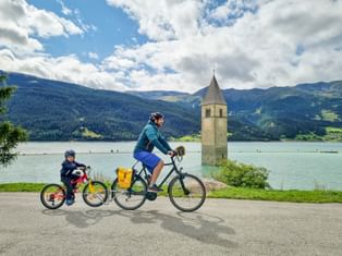 Erwachsener und Kind radeln am Uferweg des Reschensees mit dem berühmten versunkenen Kirchturm im türkisfarbenen Wasser, Berge im Hintergrund.