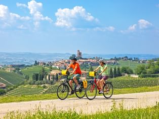 Two cyclists riding through Piedmont vineyards with Montforte village and tower visible on hilltop. Rolling hills with green vines under blue sky.