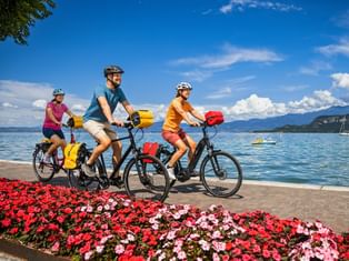 Three cyclists with panniers riding along a lakeside promenade with colorful flowers. Lake Garda and mountains visible in the background.