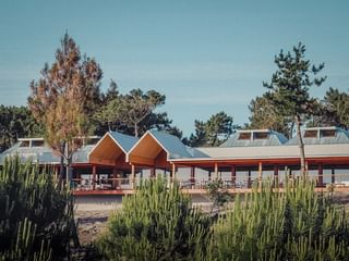 Modern hotel building with orange peaked roofs and white walls, surrounded by pine trees and green vegetation under a clear blue sky.