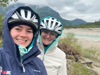 Zwei lächelnde Radfahrer mit Helmen und Regenjacken machen ein Selfie an der Isar mit Bergen und Wald im Hintergrund.