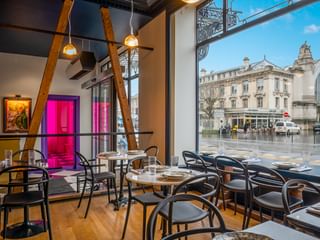 Contemporary bar interior with black bistro chairs, round tables, exposed wooden beams, and floor-to-ceiling windows overlooking a Tours street.