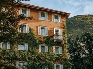 Orange four-story building with white shutters and ivy-covered walls, balcony on second floor, surrounded by trees with mountains in background.