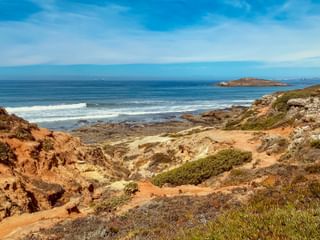 Zerklüftete Küstenlandschaft bei Porto Covo mit rotbraunen Klippen, grüner Vegetation und blauem Atlantik mit weißen Wellen und kleinen Inseln.