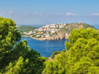 White hillside village overlooking blue Mediterranean bay in Catalonia, framed by green pine trees with distant mountains under clear sky.