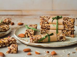 Homemade muesli bars with nuts, oats, and seeds arranged on ceramic plates. Some bars are wrapped in paper with green ribbons.