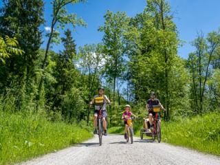 Vierköpfige Familie radelt auf Schotterweg am Lechradweg. Zwei Erwachsene und zwei Kinder mit Fahrradanhänger fahren durch grüne Landschaft unter blauem Himmel.