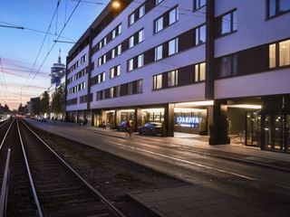 Moderne weiße Fassade des The Dean Munich Hotels bei Dämmerung mit beleuchteten Eingang und Fenstern. Straßenbahngleise verlaufen entlang der Straße.